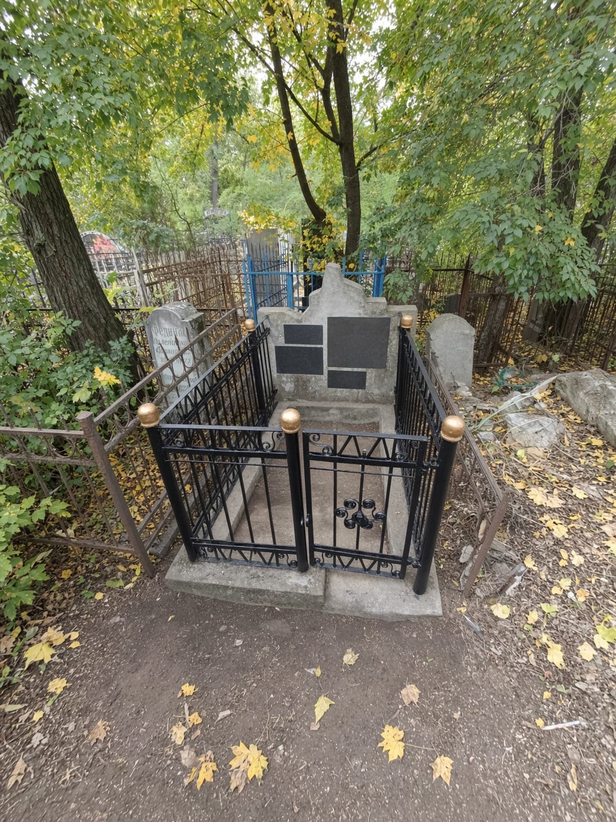 Fenced grave with grey headstone and black plaques in a wooded cemetery with autumn leaves.